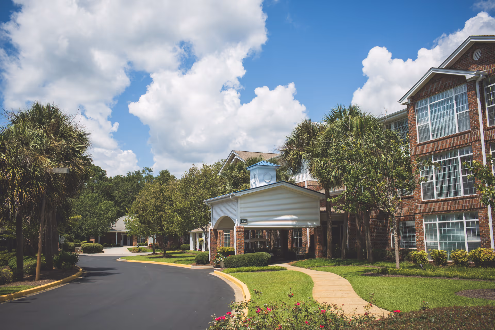 Driveway and landscaped entrance with a covered porte-cochère leading to a multi-story brick senior living building under a blue sky.