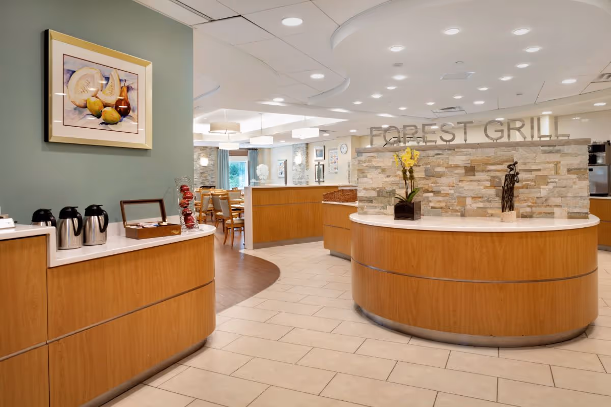 Interior view of a dining area named Forest Grill with wooden counters, a stone accent wall, and a seating area with tables and chairs in the background. The space is well-lit with ceiling lights and decorated with framed artwork and a flower arrangement on the counter.