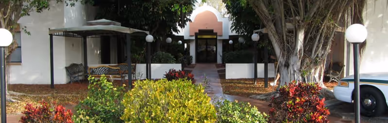 Front entrance of a building with a walkway flanked by shrubs, benches, globe lamp posts, and trees.