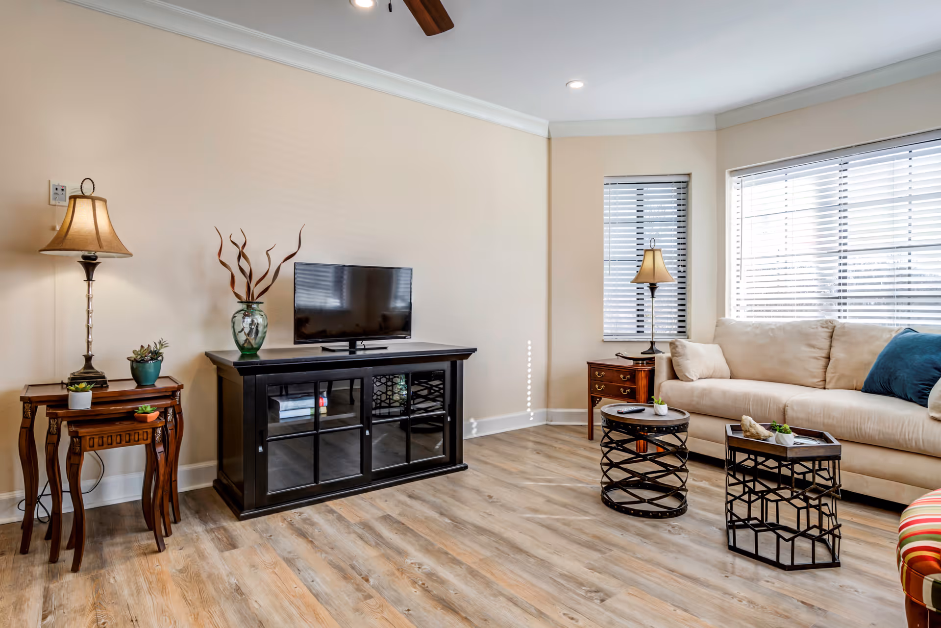 A bright living room with light-colored walls and wood flooring. The room features a beige sofa with blue and beige cushions, two round black metal coffee tables with decorative items, a black TV stand with a flat-screen TV, and two wooden side tables with lamps and small plants. Large windows with white blinds allow natural light to fill the space.