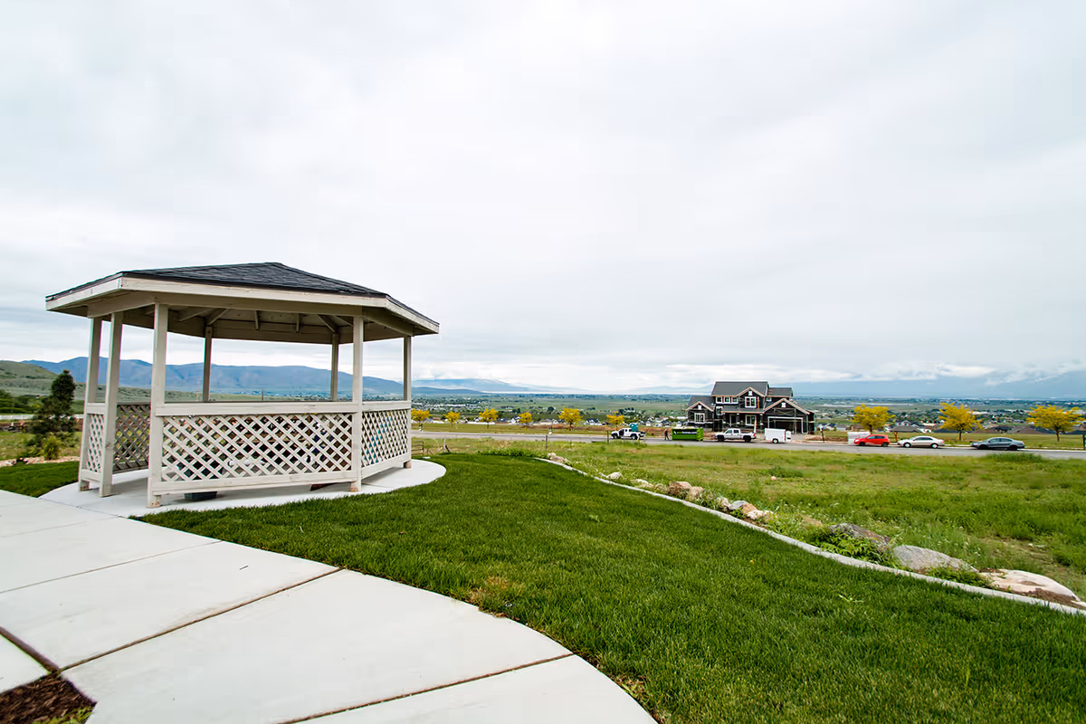 A white wooden gazebo on a concrete path surrounded by green grass with a scenic view of distant mountains and a house across the road under a cloudy sky.