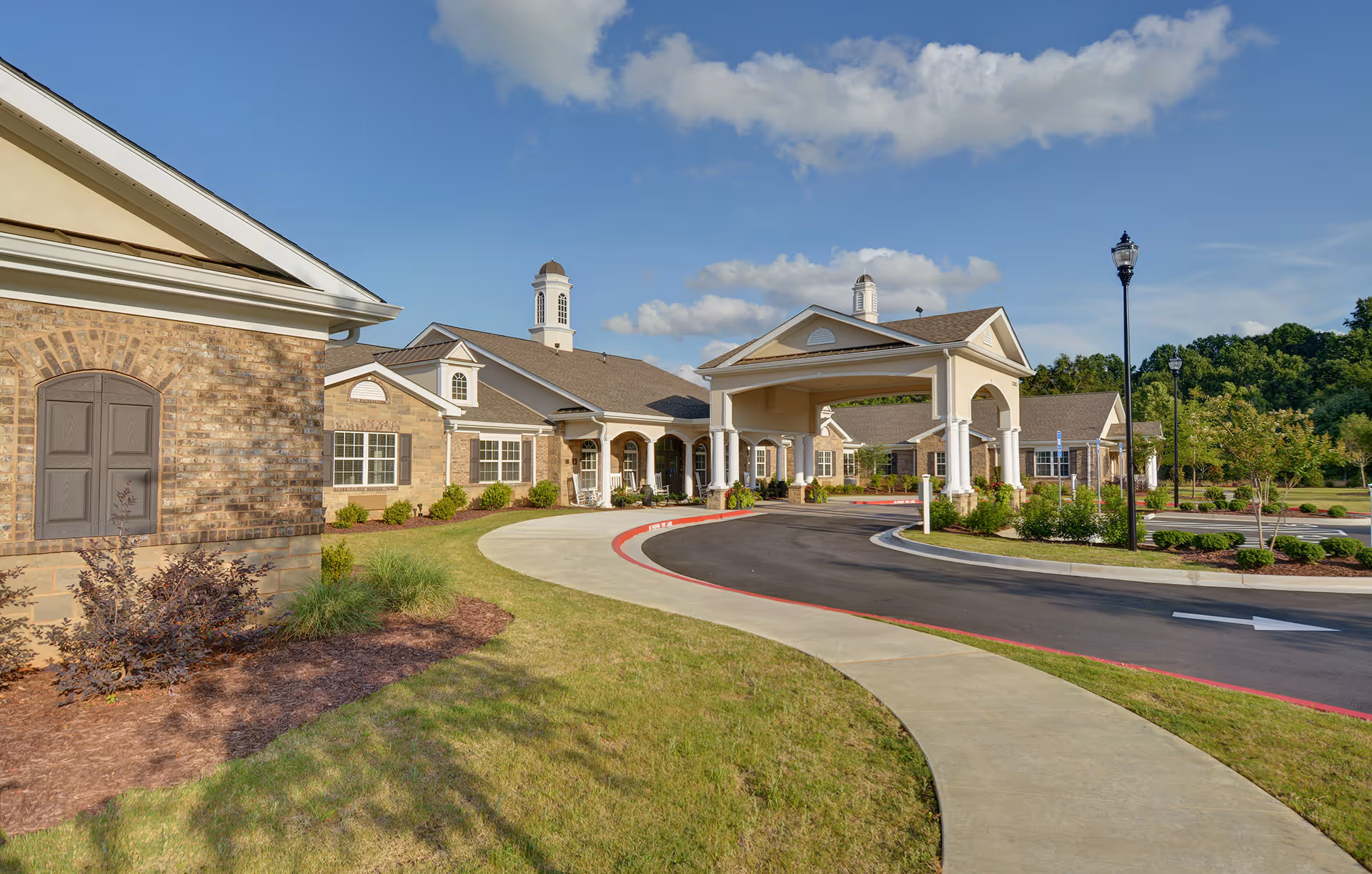 Front entrance of a brick senior living facility with a covered porte-cochère, circular driveway, and landscaped grounds under a blue sky.