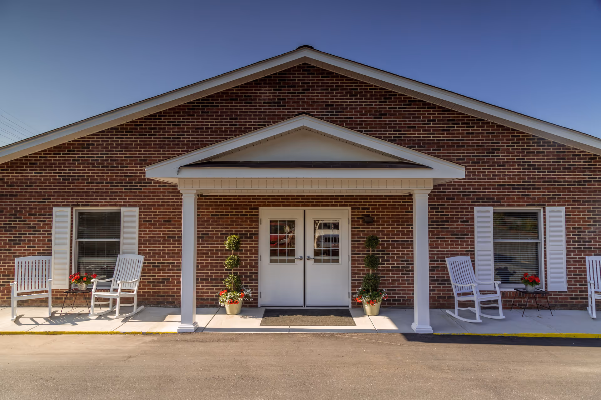 Brick building entrance with white double doors under a small portico, flanked by rocking chairs and potted plants.