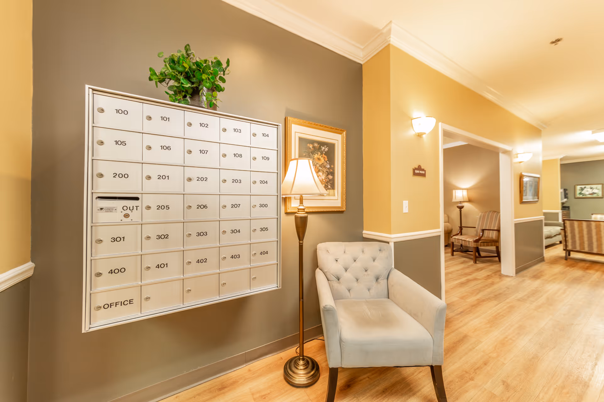 A bright assisted living lobby interior showing a wall of resident mailboxes, a potted plant, a floor lamp, an upholstered chair, and a hallway with seating.