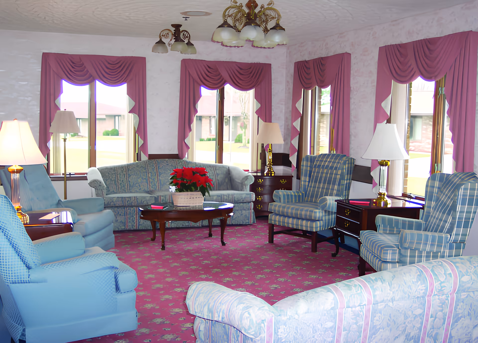 A cozy living room with multiple upholstered armchairs and sofas arranged around a wooden coffee table with a red poinsettia plant. The room features large windows with pink drapes, several table lamps, and a pink patterned carpet.