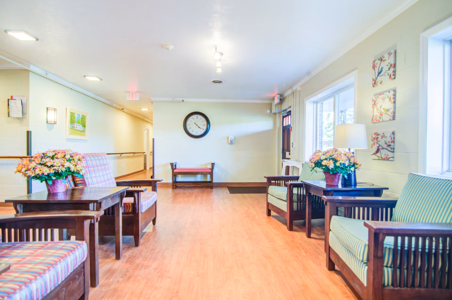 A bright and clean senior living facility common area with wooden chairs and tables arranged along the walls. The chairs have striped cushions in pastel colors. There are flower arrangements on the tables, a clock on the far wall, and three framed pictures of birds on the right wall near a window. The floor is wooden, and the walls are painted light yellow. There is a hallway leading to other rooms and an exit door at the end.