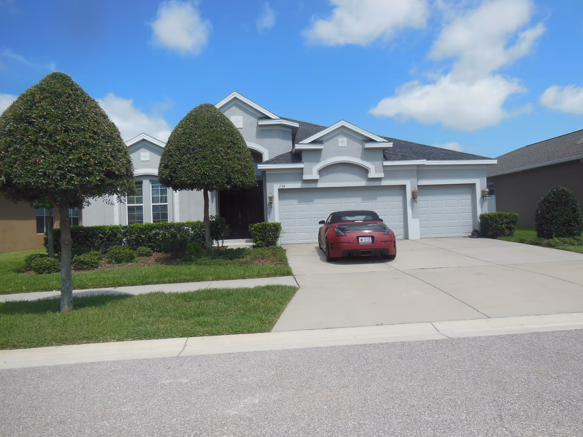 Front exterior view of a single-story residential building with a three-car garage, a red convertible car parked in the driveway, neatly trimmed bushes and trees, and a clear blue sky with some clouds.
