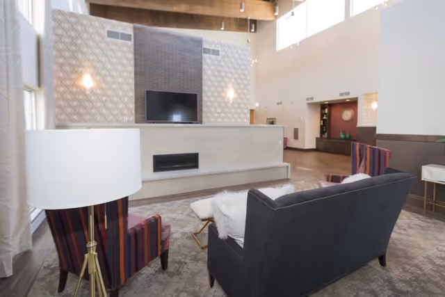 A spacious living room area with high ceilings and large windows. The room features a dark gray sofa with white pillows, two striped armchairs, a floor lamp, and a modern fireplace with a mounted flat-screen TV above it. The walls have decorative patterns and the floor is covered with a patterned rug.