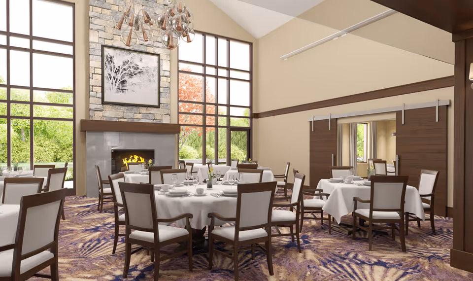 A spacious dining room with round tables covered in white tablecloths, each set with plates, glasses, and silverware. The room features large floor-to-ceiling windows letting in natural light, a stone fireplace with a fire burning, and a modern chandelier hanging from the high ceiling. The carpet has a purple and beige pattern, and there are wooden sliding doors on one side of the room.