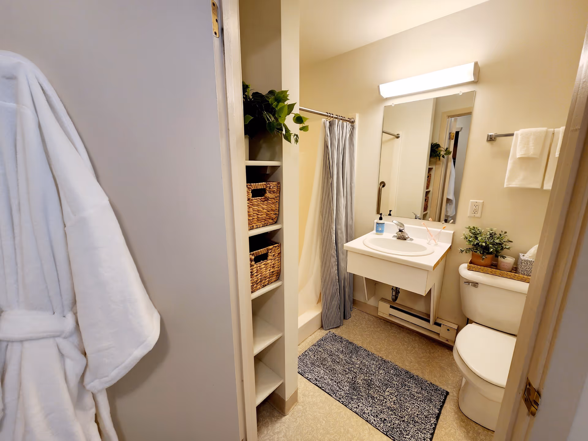 A small bathroom with a white robe hanging on the door, open shelving with woven baskets and a plant, a shower with a striped curtain, a white sink with a mirror and light above it, a soap dispenser, and a toilet with a plant and tissue box on top. A towel hangs on a rack on the wall.