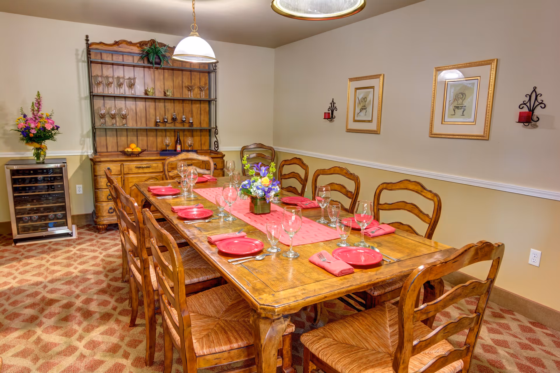 A dining room with a long wooden table set for eight people with red plates, napkins, and clear wine glasses. The room features wooden chairs, a wooden hutch with glassware and bottles, a wine cooler, a floral centerpiece on the table, and framed artwork on the walls.