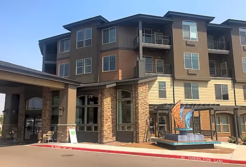 Exterior view of a multi-story senior living facility building with a stone and beige facade, large windows, balconies, and a decorative sculpture near the entrance under a clear blue sky.
