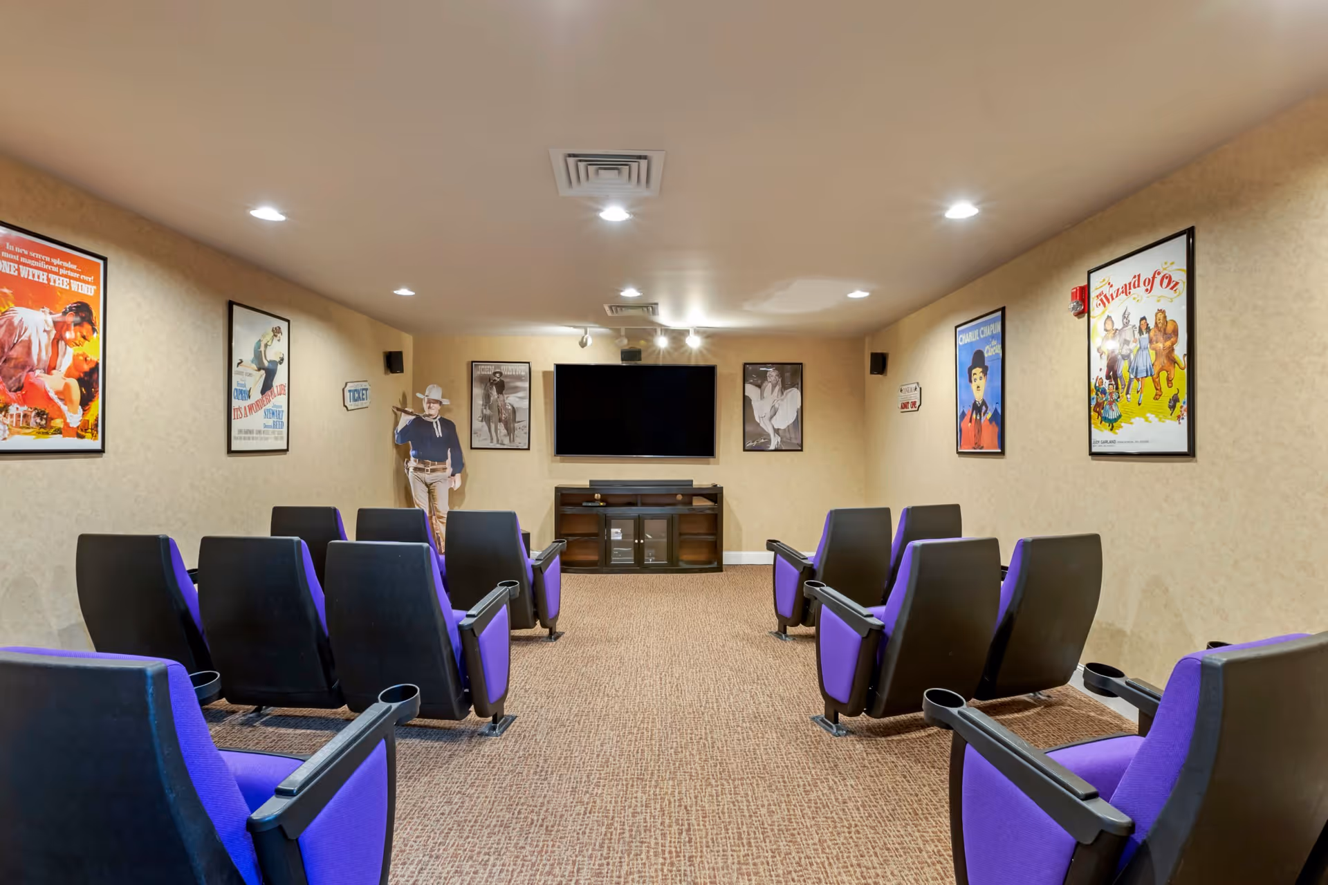 A small movie theater room with two rows of black and purple theater seats facing a large flat-screen TV mounted on a beige wall. The room is decorated with framed vintage movie posters and a cardboard cutout of a cowboy figure. The carpet is brown, and the ceiling has recessed lighting.