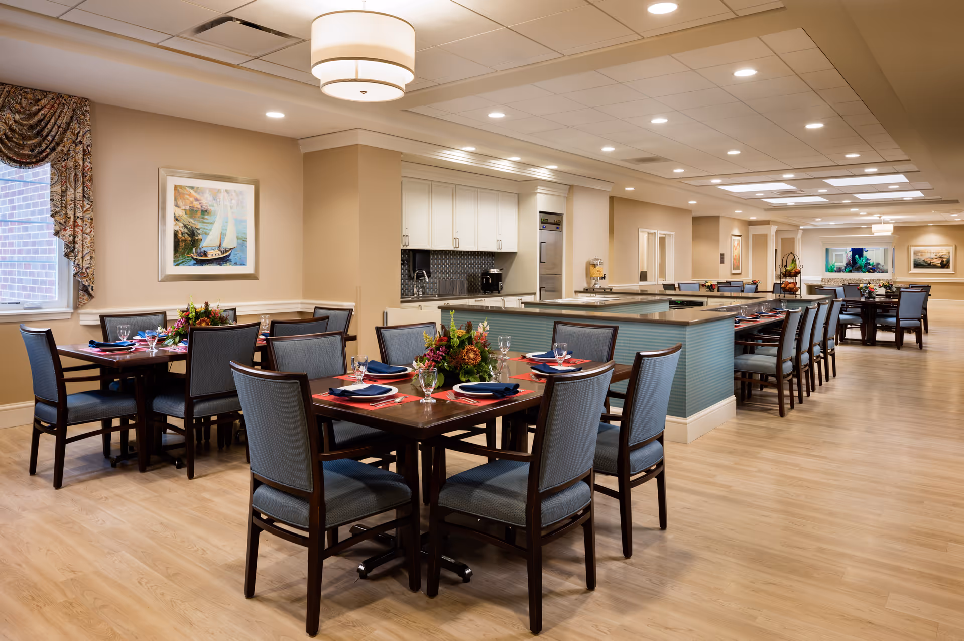 A spacious and well-lit dining room with multiple wooden tables and chairs arranged neatly. Each table is set with plates, glasses, napkins, and floral centerpieces. The room features light wood flooring, beige walls, a large window with patterned curtains, and ceiling lights. There is a kitchen area with white cabinets and appliances visible in the background.