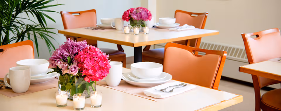 A dining area with tables set for a meal, featuring white bowls, plates, cups, and silverware on beige placemats. Each table has a centerpiece with vibrant pink and purple flowers in a glass vase, surrounded by small lit candles. Orange chairs are arranged around the tables, and a green plant is visible in the background.