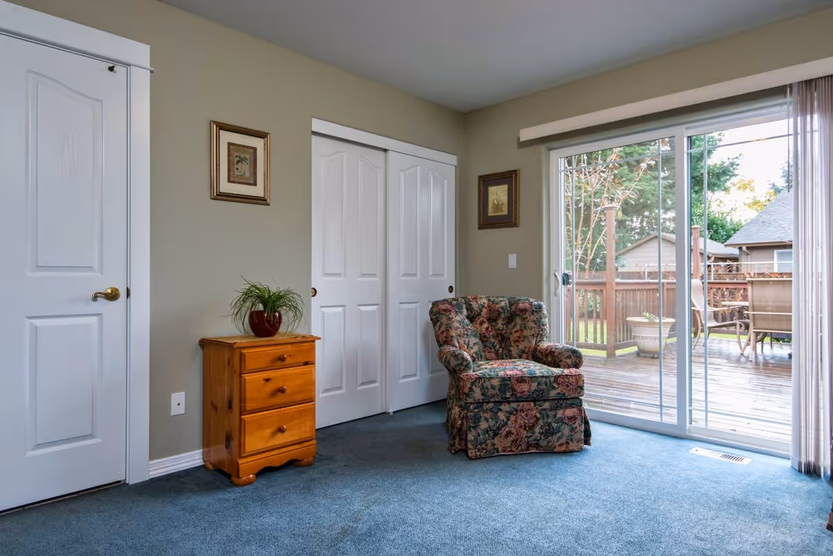 Carpeted interior room featuring a floral upholstered armchair, a small wooden dresser, closet doors, and sliding glass doors leading to a backyard deck.