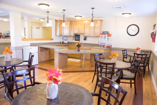 Bright communal dining area with round tables and chairs and a central buffet counter/kitchenette under pendant lights.