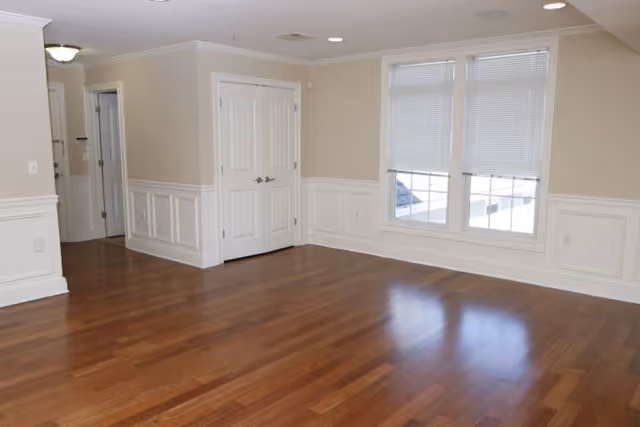 Empty room with polished hardwood floors, beige walls with white wainscoting, two white double doors, and two windows with closed blinds letting in natural light.