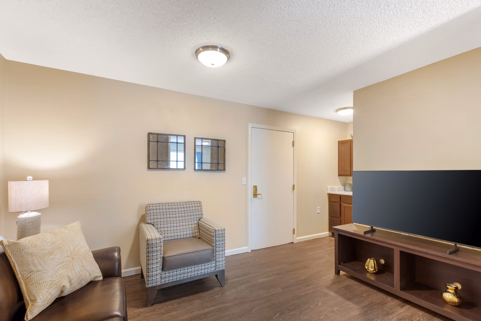 A cozy living room area with a brown leather sofa with a patterned cushion, a patterned armchair, two decorative mirrors on the beige wall, a wooden TV stand with a large flat-screen TV, and a small kitchen area visible in the background.