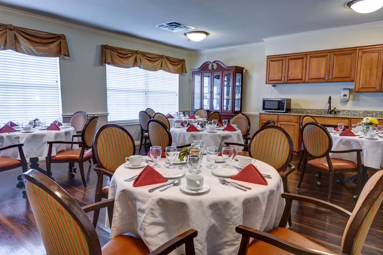A dining room with round tables covered in white tablecloths, each set with cups, glasses, silverware, and red folded napkins. The room has wooden chairs with striped upholstery, large windows with blinds and brown valances, wooden cabinets, a microwave, and a glass-fronted china cabinet.