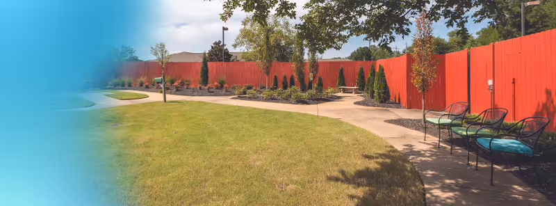 Landscaped outdoor courtyard with a curved concrete path, grassy area, benches, and a tall red wooden fence.