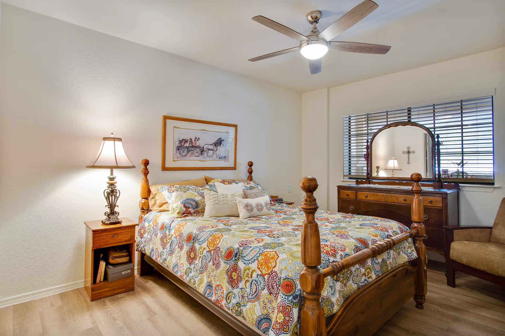 A wooden four-poster bed with colorful patterned bedding in a warmly lit bedroom featuring a nightstand with a lamp, dresser with mirror, ceiling fan, and window blinds.