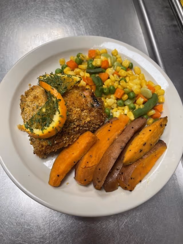 A white plate with a breaded and baked chicken breast garnished with two lemon slices and herbs, served with a side of mixed vegetables including corn, peas, green beans, and carrots, and several roasted sweet potato wedges.