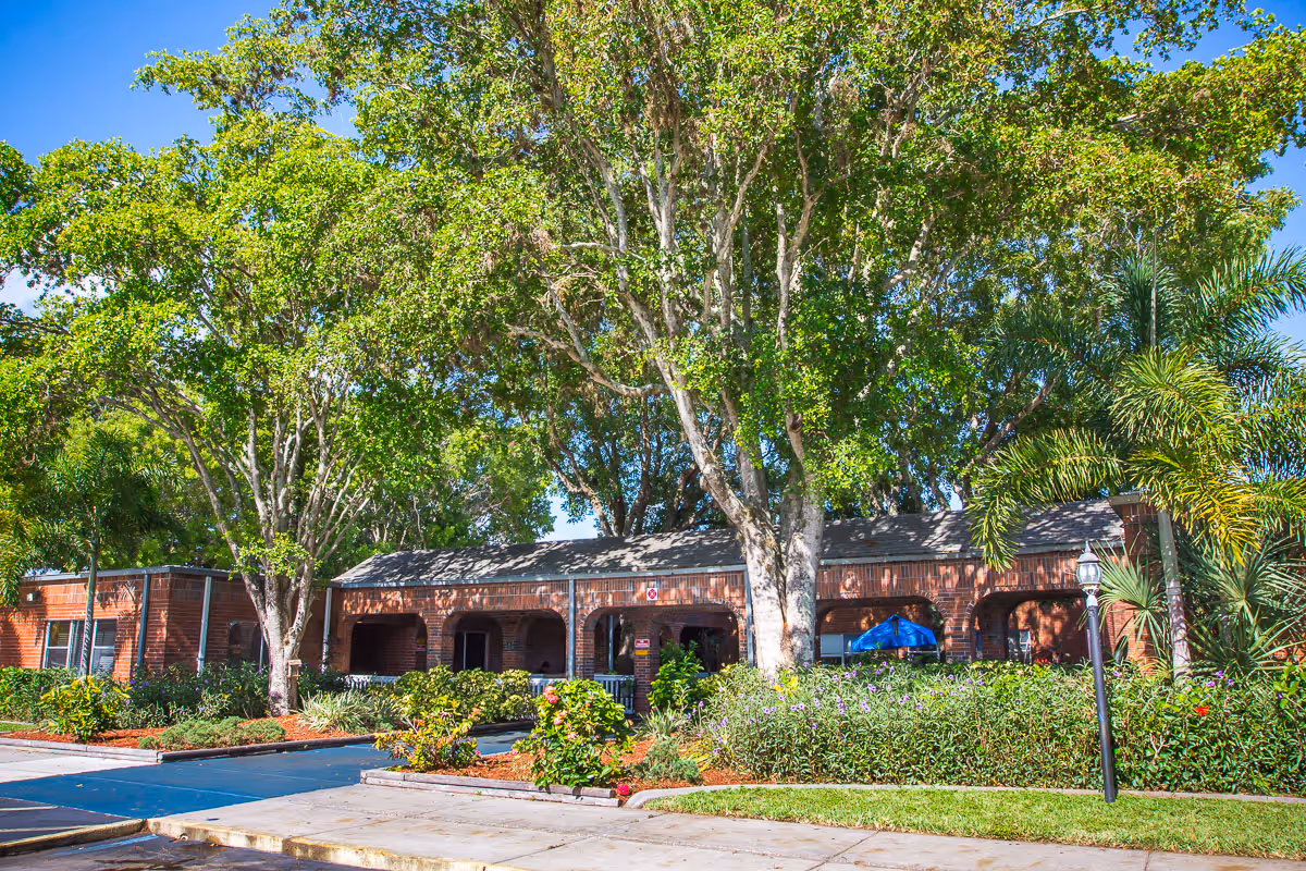 Front exterior of a single-story brick rehab and wellness center with large shade trees, landscaped beds, and a driveway.