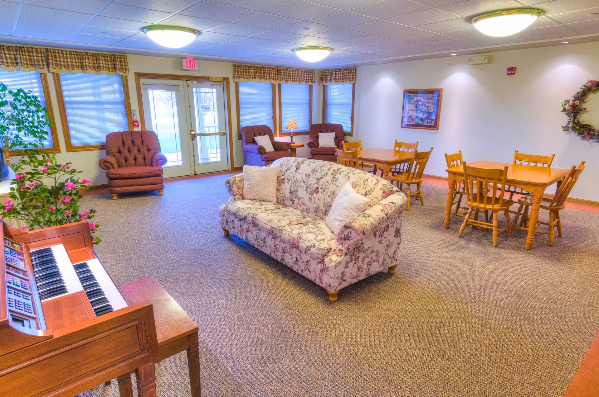 A spacious common area with a floral patterned sofa in the center, two brown armchairs near windows with plaid valances, wooden tables and chairs arranged for seating, a wooden organ with a plant on top, and a framed picture and floral wreath on the walls.