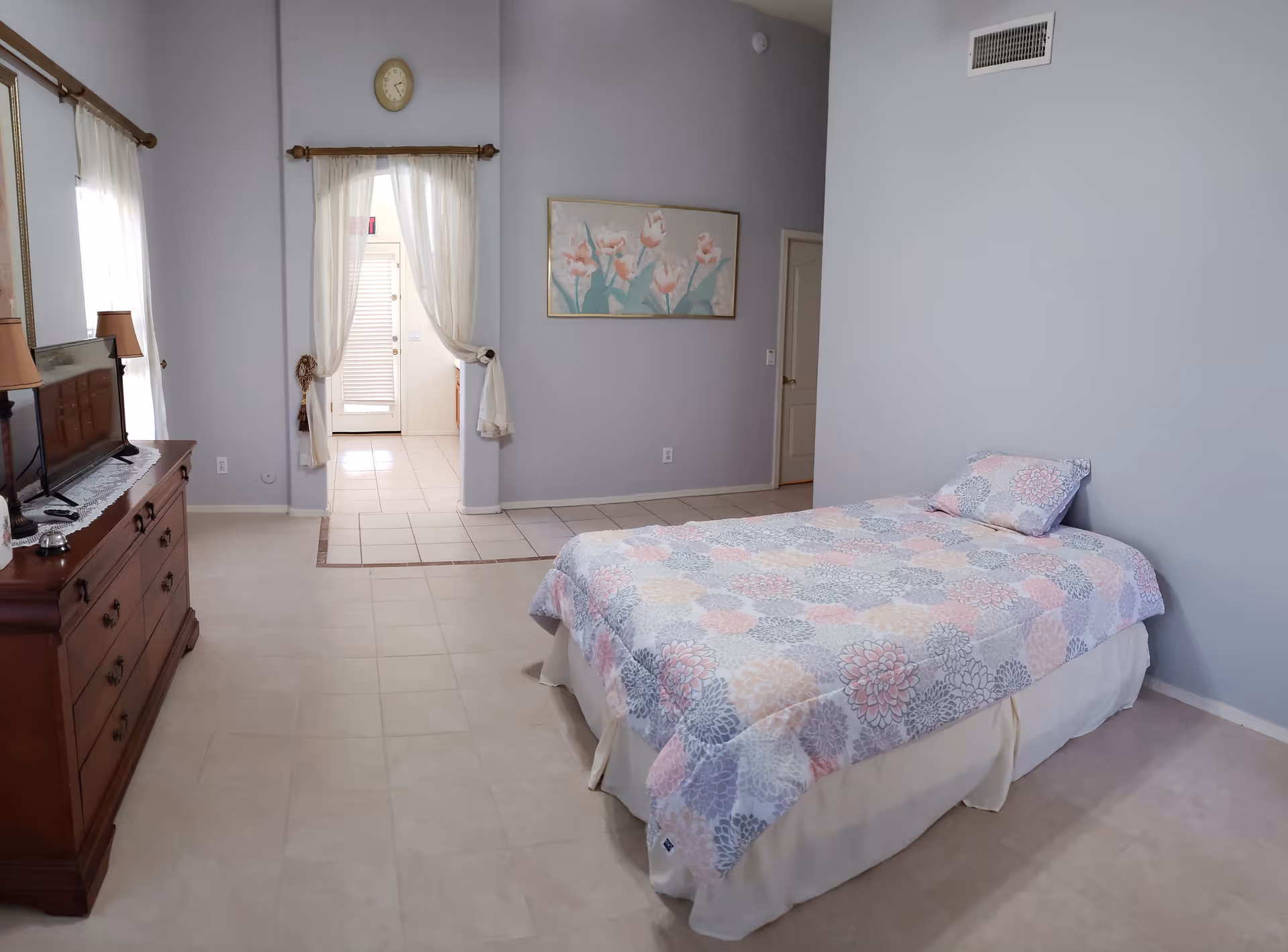 A spacious bedroom featuring a single bed with floral bedding, a wooden dresser with a TV, and an open doorway with curtains.