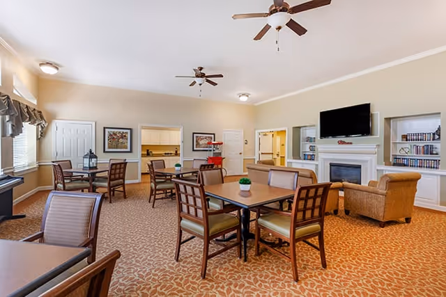 A spacious lounge area featuring several tables and chairs, a fireplace, and a television. The room has light-colored walls and large windows allowing natural light to enter.