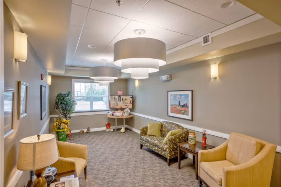A cozy sitting area in a senior living facility with two yellow armchairs, a patterned loveseat with green pillows, a small wooden side table, and a lamp. The room has beige walls, carpeted floor, modern ceiling lights, framed artwork on the wall, and a window with natural light. There is also a corner table with various items and a potted plant near the window.