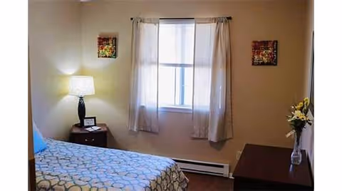 A bedroom with a bed covered in a blue and white patterned bedspread, a nightstand with a lamp and a small picture frame, a window with white curtains, two framed pictures on the walls, and a table with a vase of flowers.