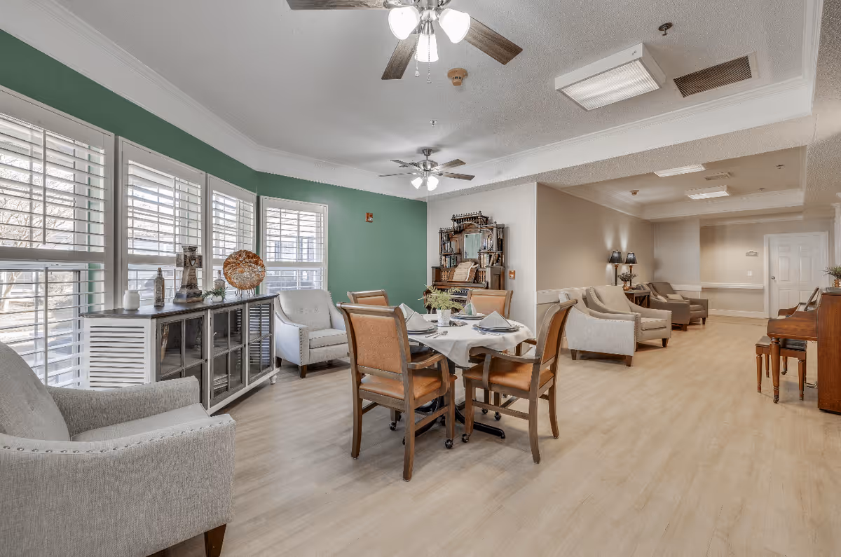 A bright and spacious common area in a senior living facility with large windows featuring white shutters, a green accent wall, and light wood flooring. The room is furnished with a dining table set for four, several upholstered armchairs, a wooden piano, and decorative items on a sideboard. Ceiling fans with lights hang from the ceiling.