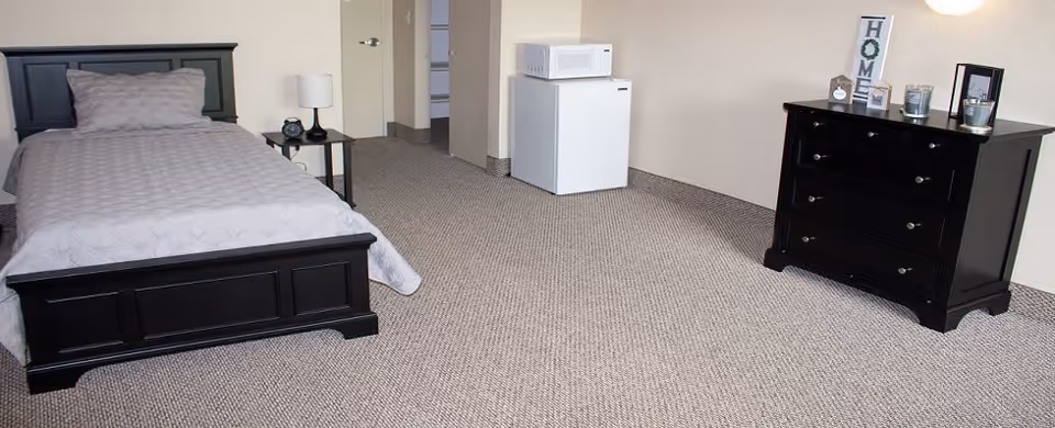 A simple bedroom with a single bed with gray bedding, a small black nightstand with a lamp and clock, a white mini refrigerator with a microwave on top, and a black dresser with decorative items including a framed 'HOME' sign and candles.
