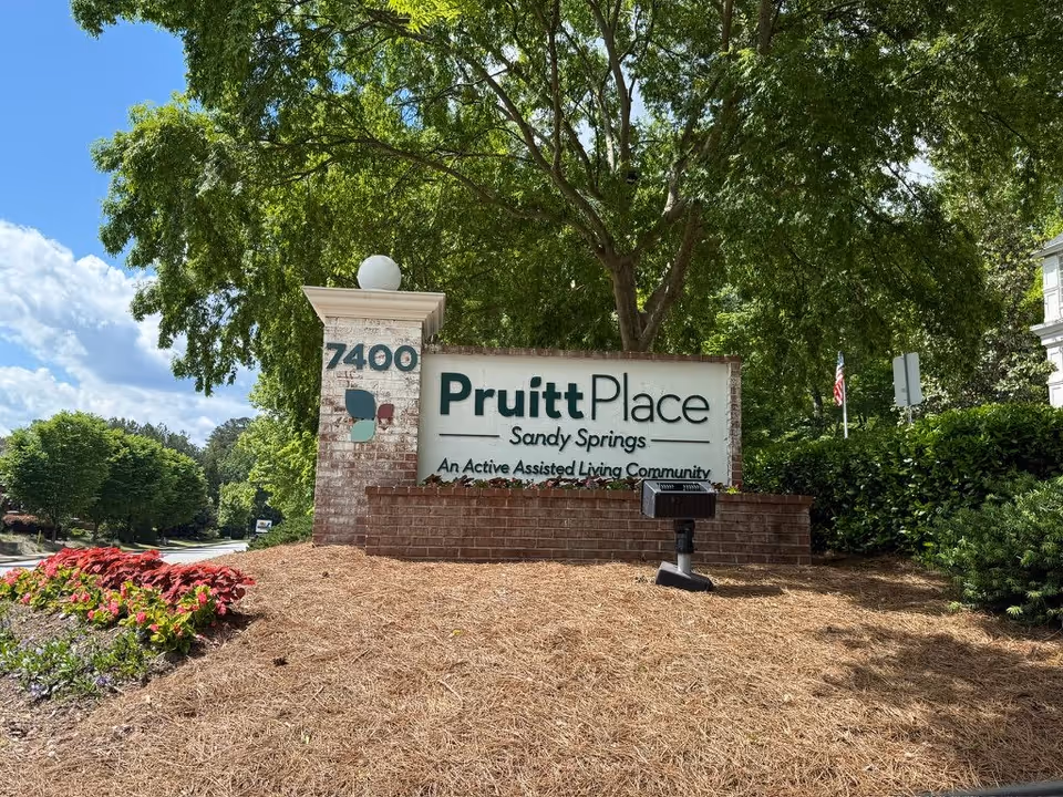 Brick entrance sign reading 'PruittPlace Sandy Springs' at 7400 surrounded by landscaping and trees under a blue sky.