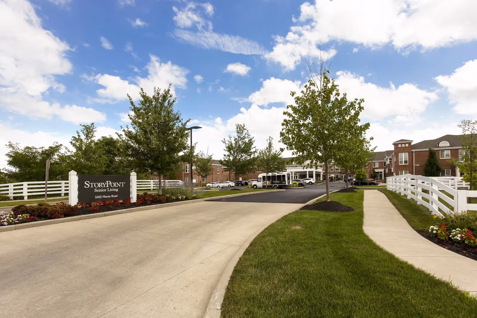 Entrance driveway and sidewalk leading to the StoryPoint Senior Living facility building, with neatly maintained green lawns, trees, flower beds, and a white fence under a partly cloudy blue sky.