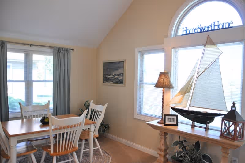 A bright dining area with a wooden table and four white chairs with wooden seats. There are two windows with blinds and curtains, a small table with a lamp, a decorative sailboat, a lantern, and a framed photo. Above the window is a decorative sign that reads 'Home Sweet Home'.