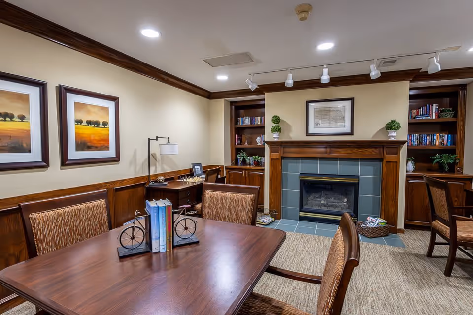 A cozy interior room with a wooden table and four chairs, a fireplace with a wooden mantle, built-in bookshelves filled with books and plants, two framed landscape paintings on the wall, and a small desk with a lamp and chessboard in the corner.
