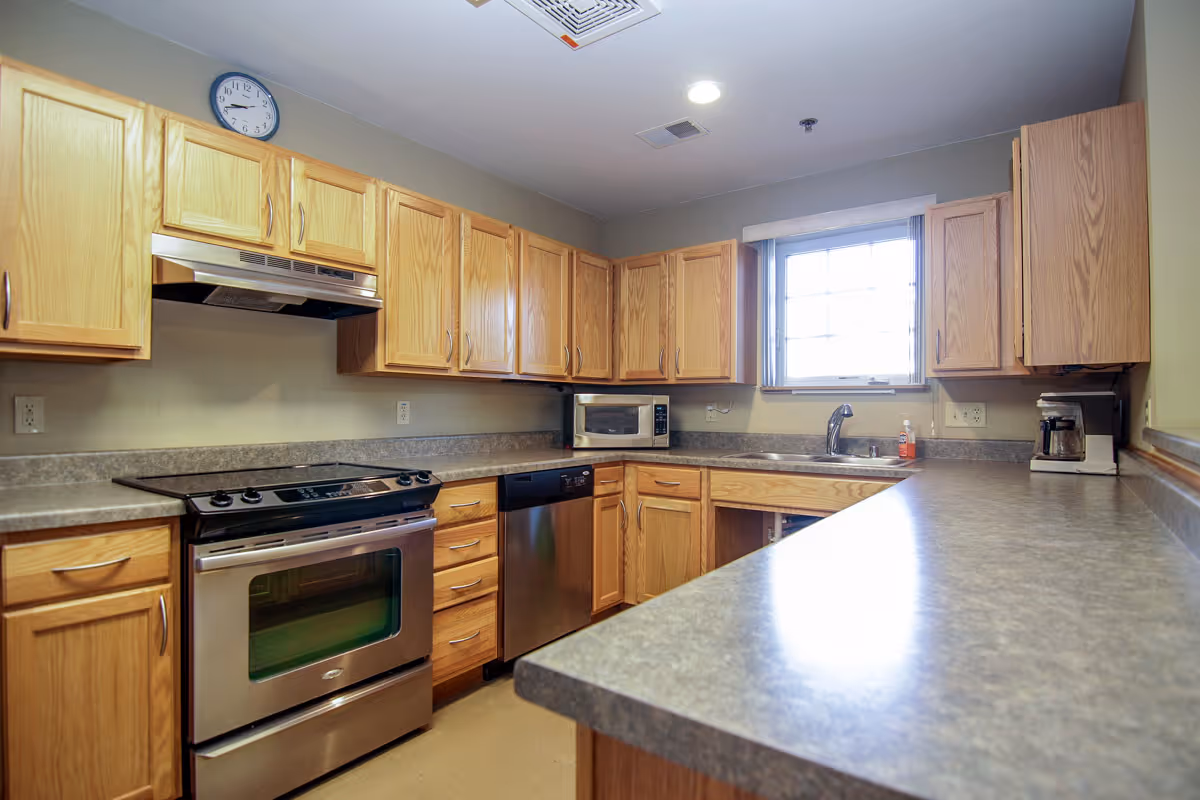 A kitchen with wooden cabinets, a stainless steel oven and dishwasher, a microwave, a coffee maker, a sink under a window, and a long countertop.