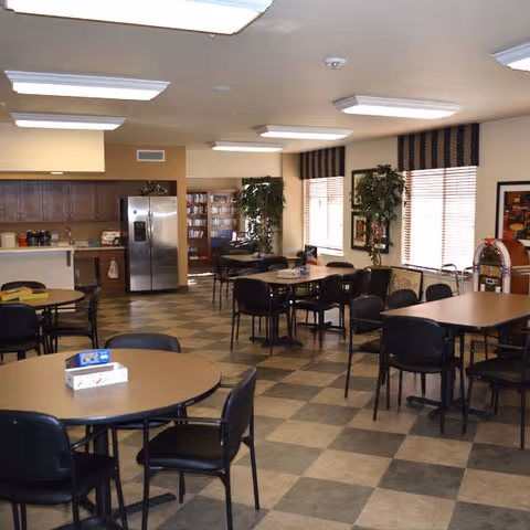 Large communal dining/activity room with multiple round and rectangular tables and chairs, a kitchenette with refrigerator in the back, and windows with blinds.