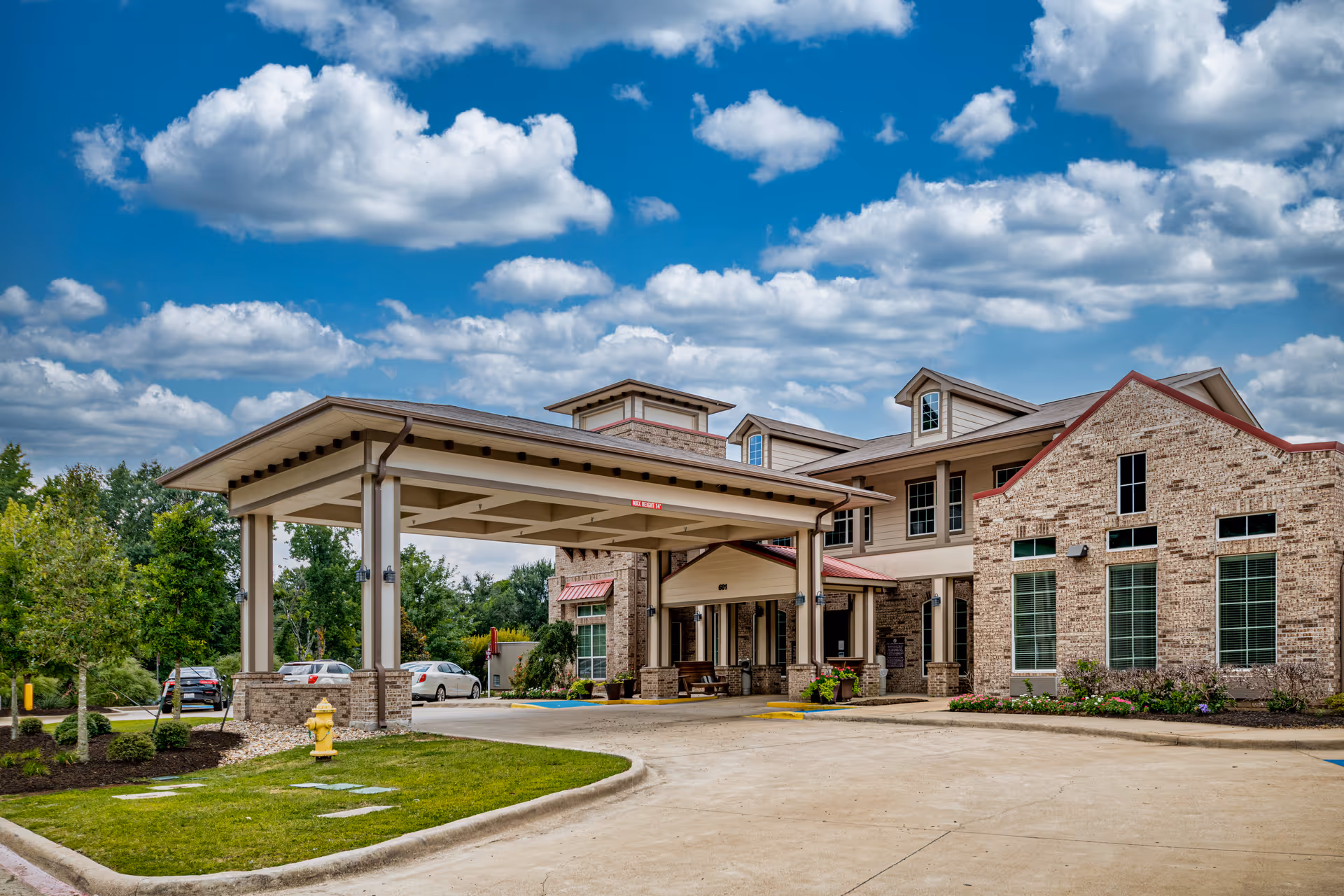Exterior view of a senior living facility named Parkview on Hollybrook, featuring a covered entrance with a driveway, brick building with multiple windows, and a partly cloudy blue sky.