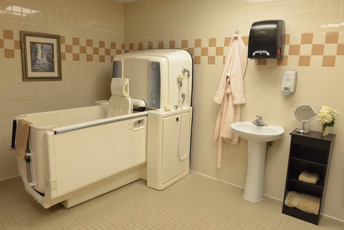 A bathroom with beige tiled walls and floor, featuring a walk-in bathtub with a seat and handrails, a pedestal sink with a faucet, a wall-mounted soap dispenser, a black paper towel dispenser, a pink bathrobe hanging on the wall, a small black shelving unit with towels and a vase of flowers, and a framed picture on the wall.