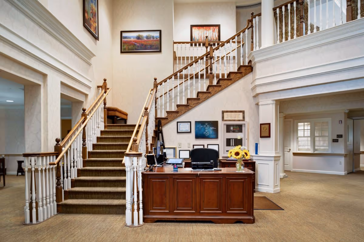 Reception desk with a vase of sunflowers in front of a grand double staircase and framed artwork in a senior living facility lobby.