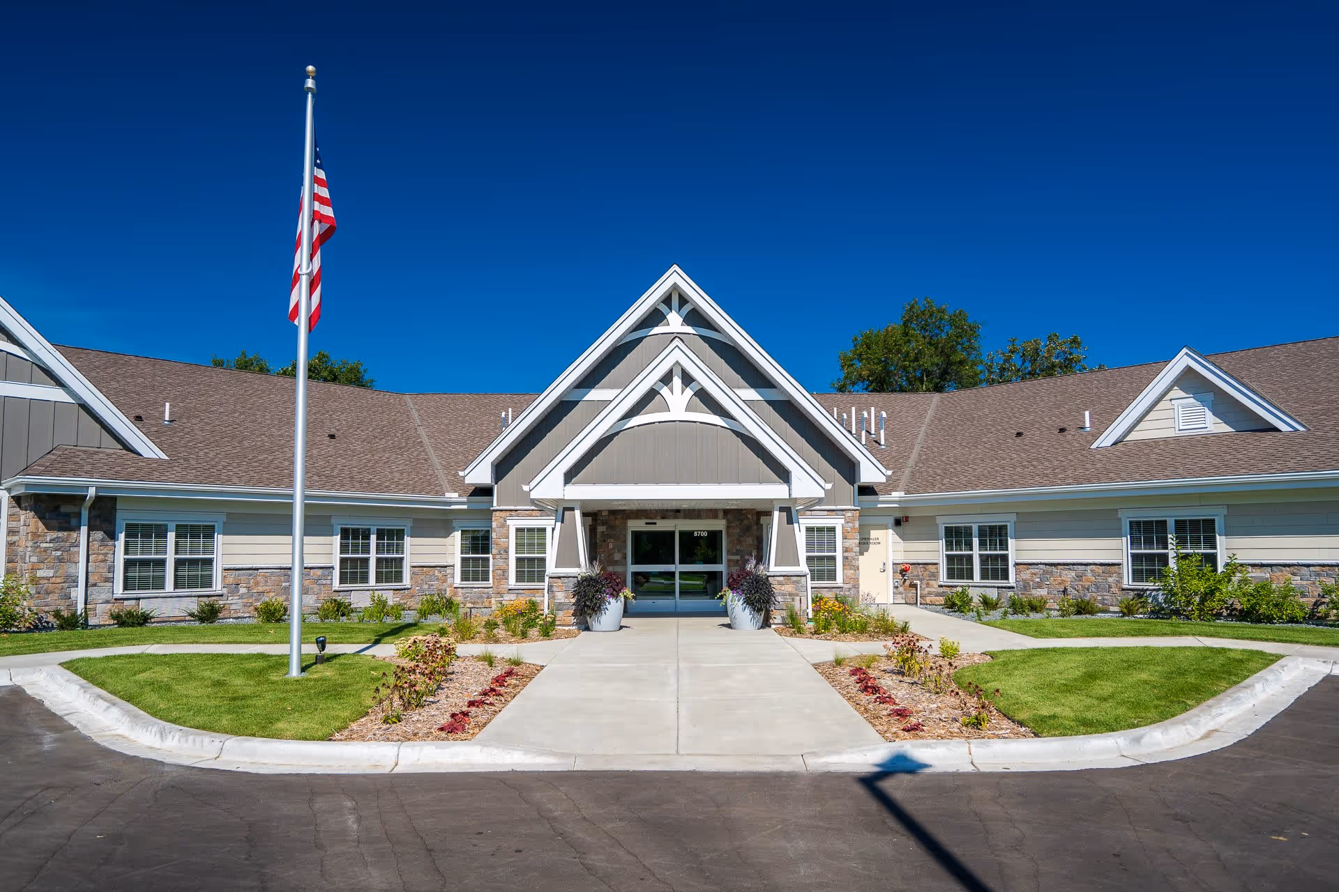 Front entrance of a single-story senior living building with an American flag, paved driveway, and landscaped walkway under a clear blue sky.