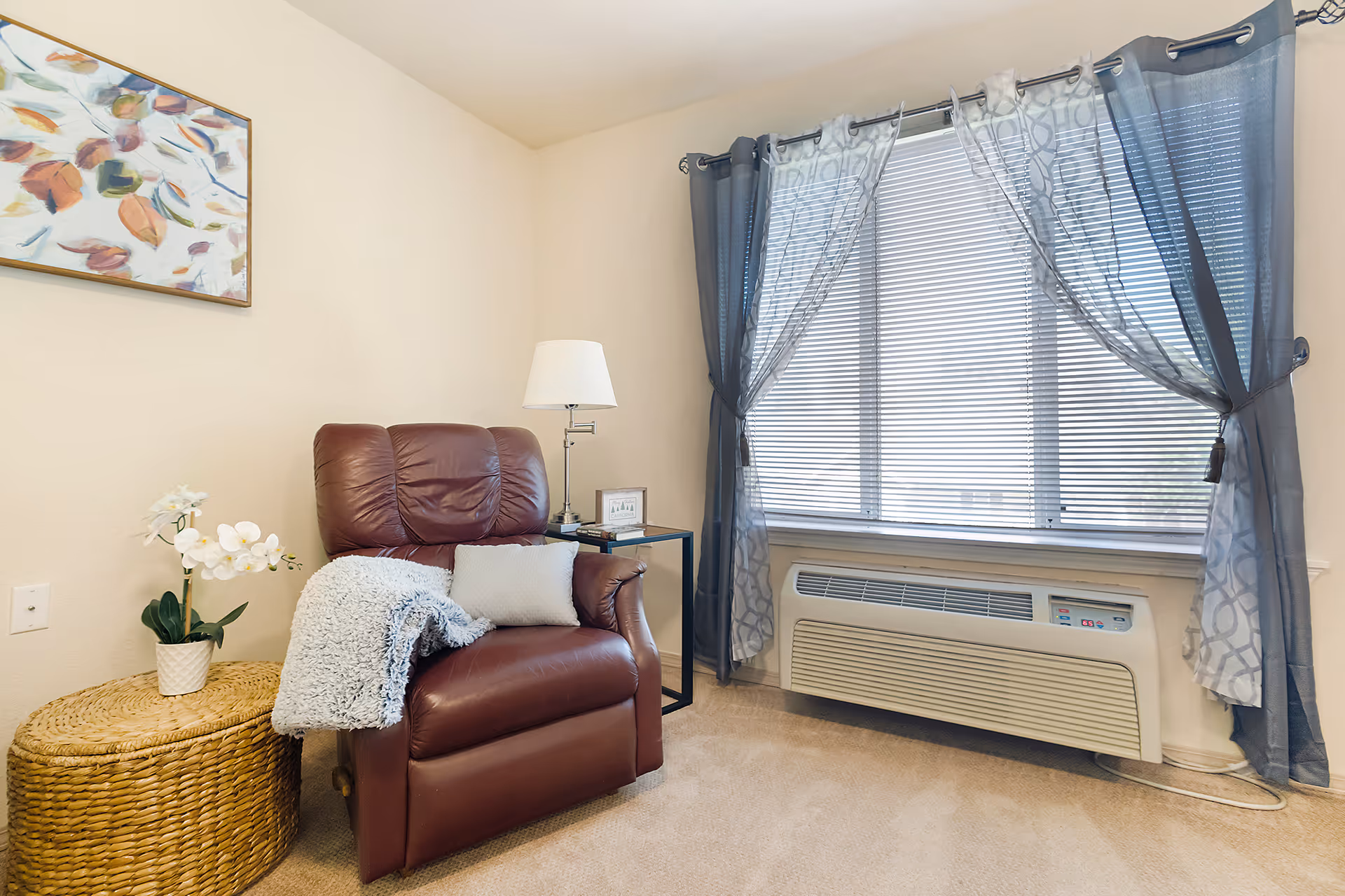 A cozy corner of a room featuring a brown leather recliner chair with a gray throw blanket and a light-colored pillow. Next to the chair is a round wicker side table with a white orchid plant on it. A small black side table with a lamp and a framed picture is positioned beside the chair. The window behind has closed blinds and sheer gray curtains tied back on each side. The room has beige walls and carpeted floor.