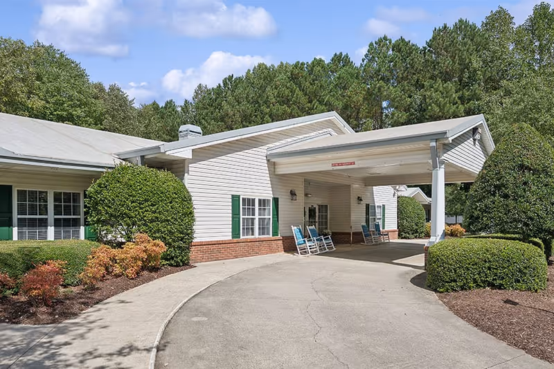 Exterior view of a single-story building with white siding and green shutters, featuring a covered entrance with a driveway. There are blue rocking chairs under the covered area and well-maintained bushes and trees surrounding the building.
