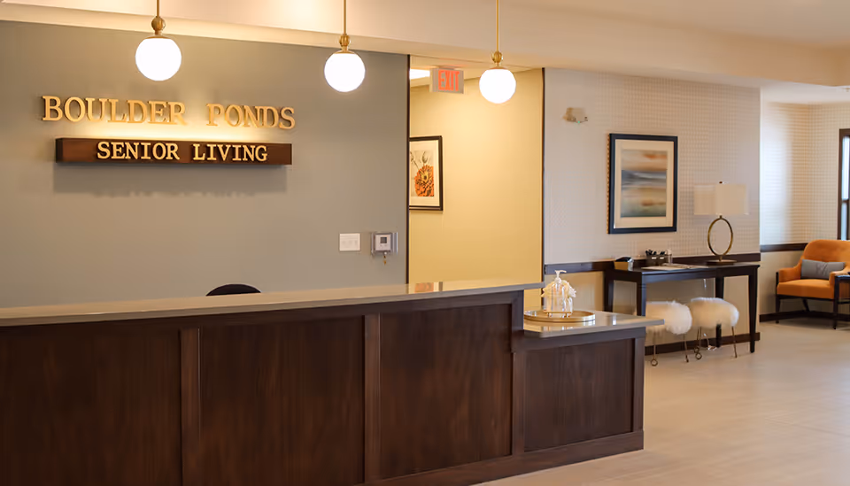 Reception desk and lobby of Boulder Ponds Senior Living with a wooden front desk, wall sign, pendant lights and seating area.