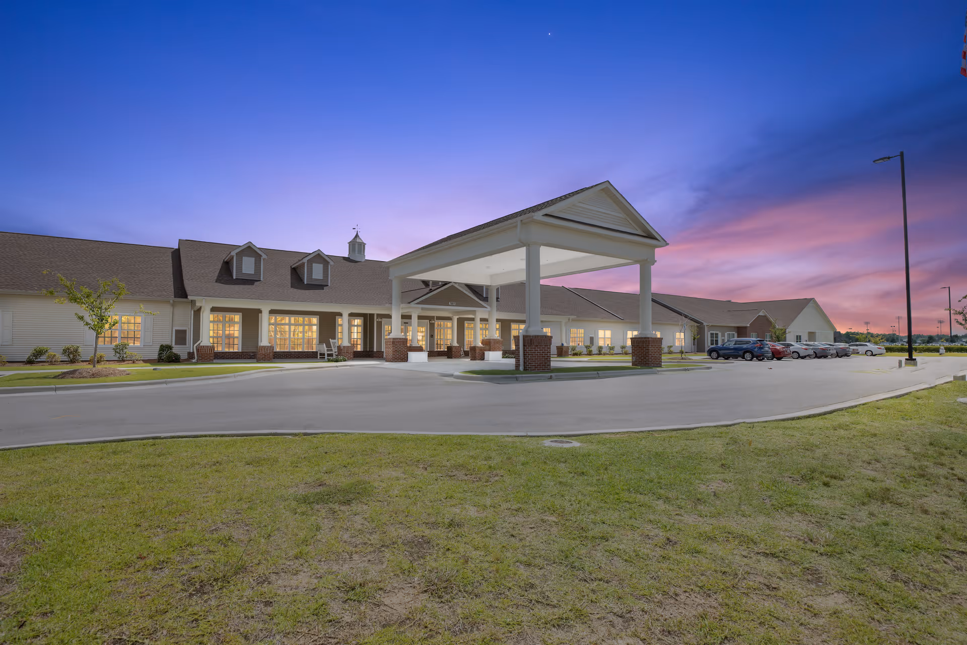 Front exterior of a senior living building with a covered entrance and parking lot at dusk.