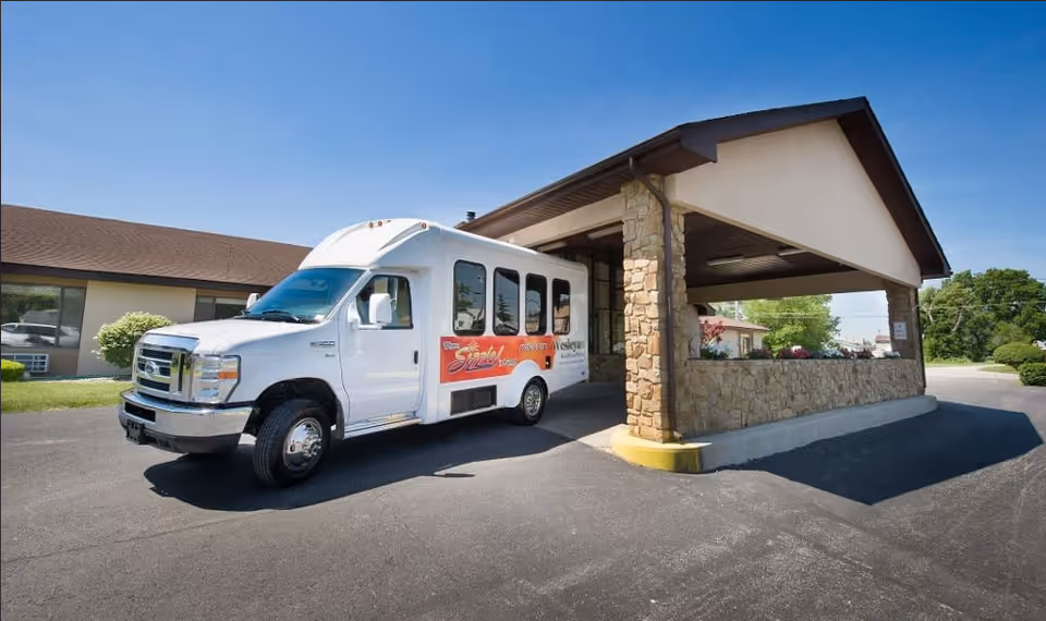 A white shuttle van parked under a covered entrance of a single-story building with stone pillars and beige walls, surrounded by greenery and a clear blue sky.